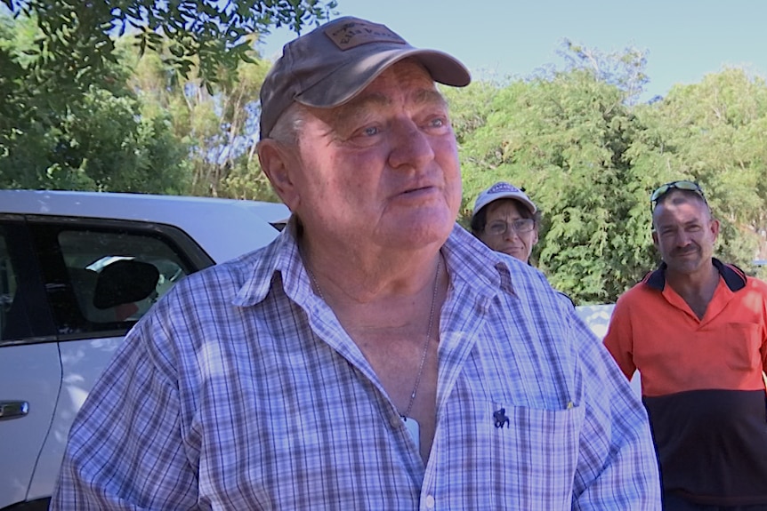 An older man wearing a cap stands outside in front of a white vehicle with two other men in the background.