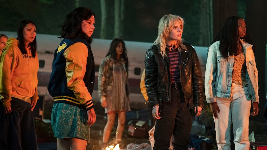 A photo shows a group of young women dressed in jackets standing around a camp fire