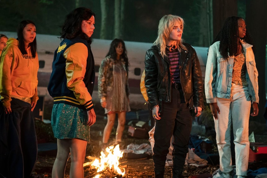 A photo shows a group of young women dressed in jackets standing around a camp fire