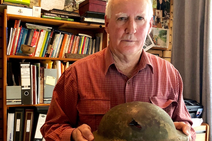 John Tannock stands in his house holding a helmet worn by his father during WWII.