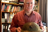 John Tannock stands in his house holding a helmet worn by his father during WWII.