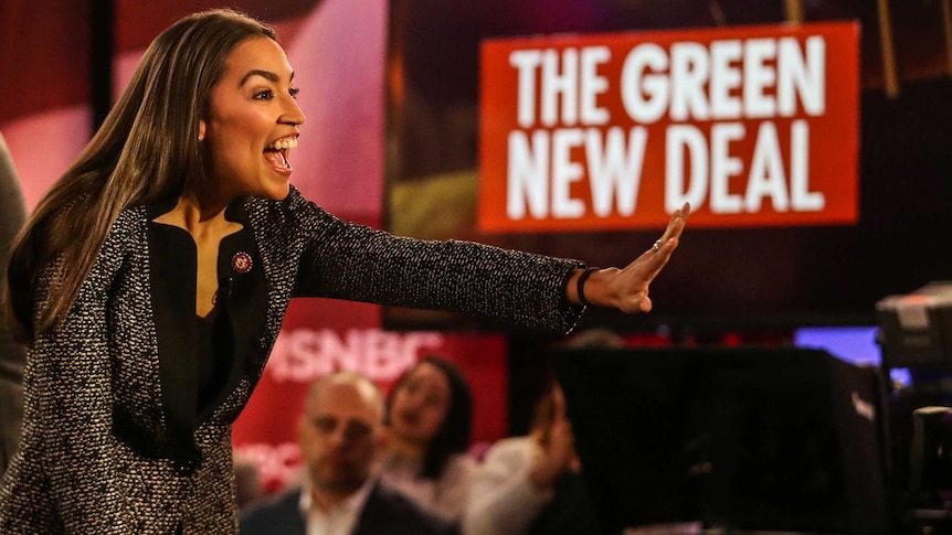 Alexandria Ocasio Cortez waves and smiles to a crowd as a sign bearing the words "Green New Deal" is visible in the background.