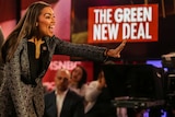 Alexandria Ocasio Cortez waves and smiles to a crowd as a sign bearing the words "Green New Deal" is visible in the background.