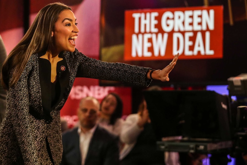 Alexandria Ocasio Cortez waves and smiles to a crowd as a sign bearing the words "Green New Deal" is visible in the background.