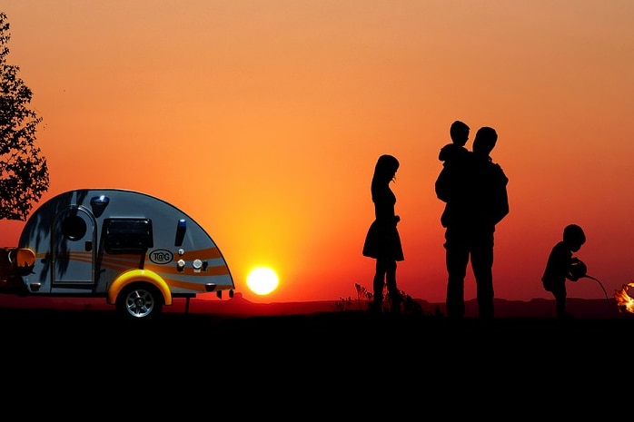 A family silhouetted at sunset with a campfire and a caravan.