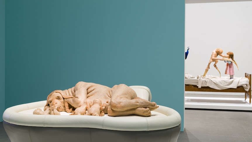 Life-sized sculpture of a genetically-engineered dog/human hybrid nursing a litter of pups, within a gallery with blue walls.