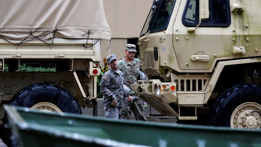 Two men in camouflage stand near military vehicles.