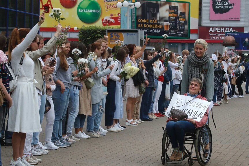 A woman pushes a woman in a wheelchair past a long chain of women holding white flowers aloft, waving hands and smiling.