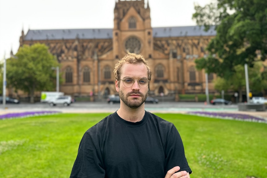 A man wearing glasses and a black tshirt stands with his arms crossed