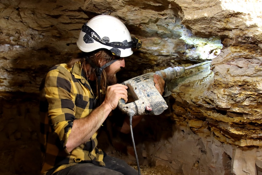 A man with long hair wearing a hardhat holds a handheld jackhammer into the rock wall of a mine. 