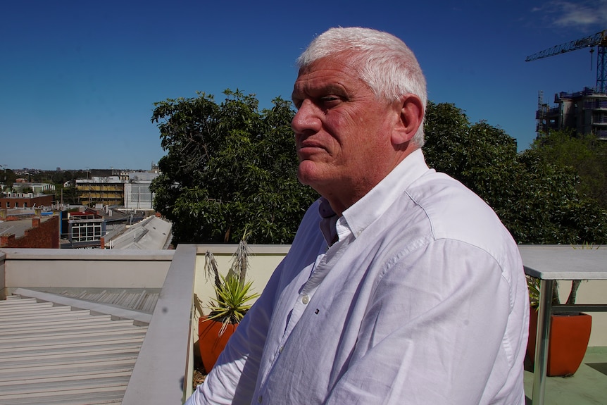 A man stands on a rooftop, looking to the side of frame with a serious expression.