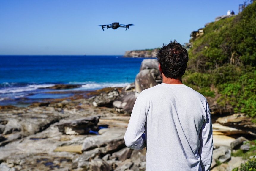 A man stands with his back to the camera looking at a beach, operating a drone which can be seen in the sky,