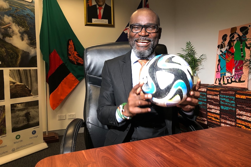 A man, sitting at a desk, holds a soccer ball and has a wide grin. 
