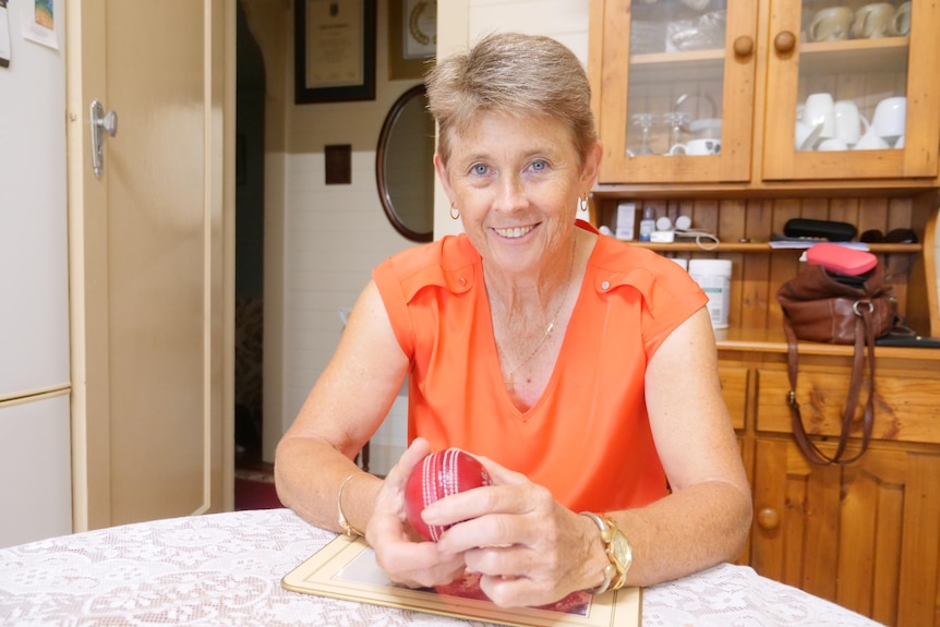 A woman sits at a table holding a cricket ball in front of her and smiling at the camera.