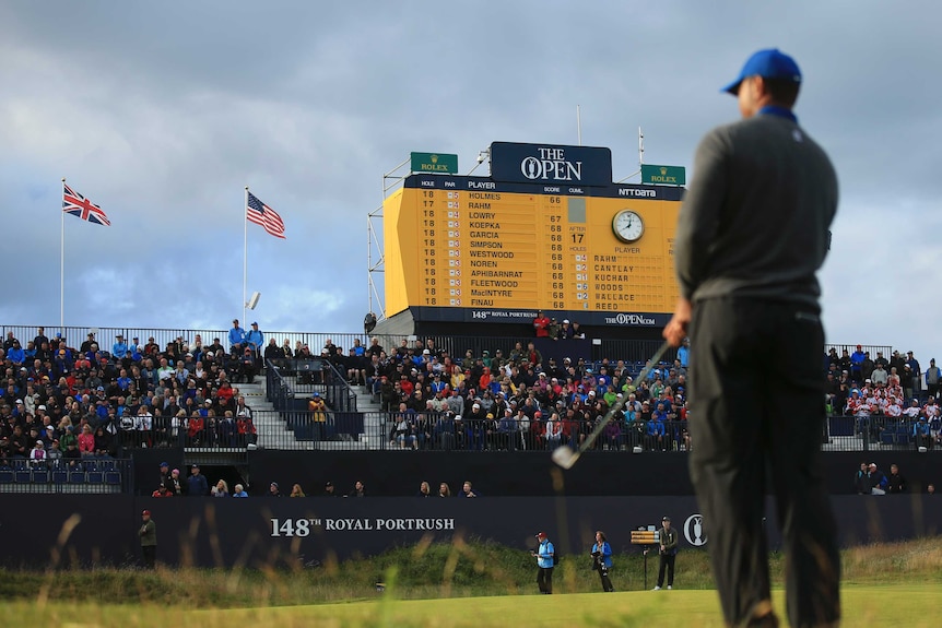 A golfer stands with his back to camera, looking at the green, with the leaderboard in front of him.