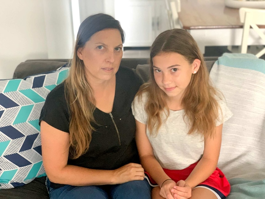 A mother and her teenage daughter look seriously at the camera, sitting inside their home.