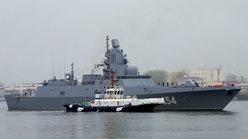 A grey metal navy warship sits in a port accompanied by a tugboat.