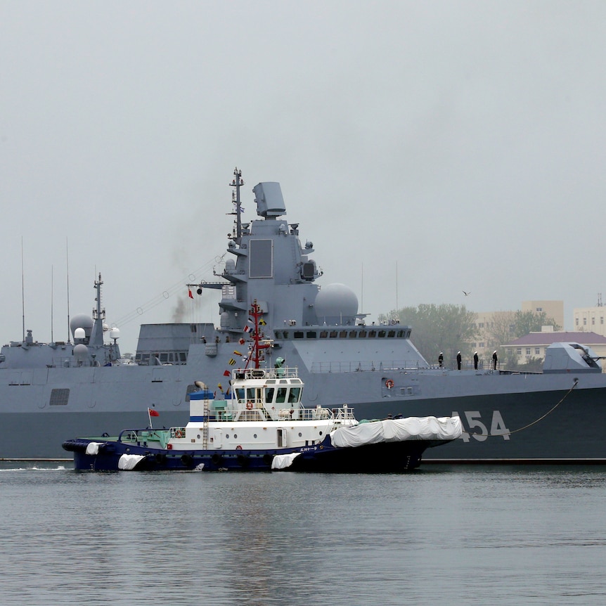 A grey metal navy warship sits in a port accompanied by a tugboat.