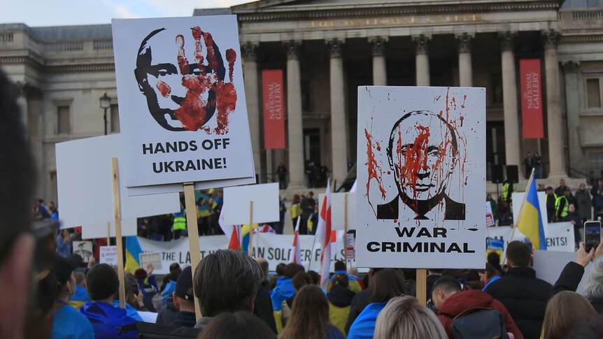 back of protestors wearing ukraine flag on their backs hold protest signs of vladimir putin with slogan "war criminal" and more