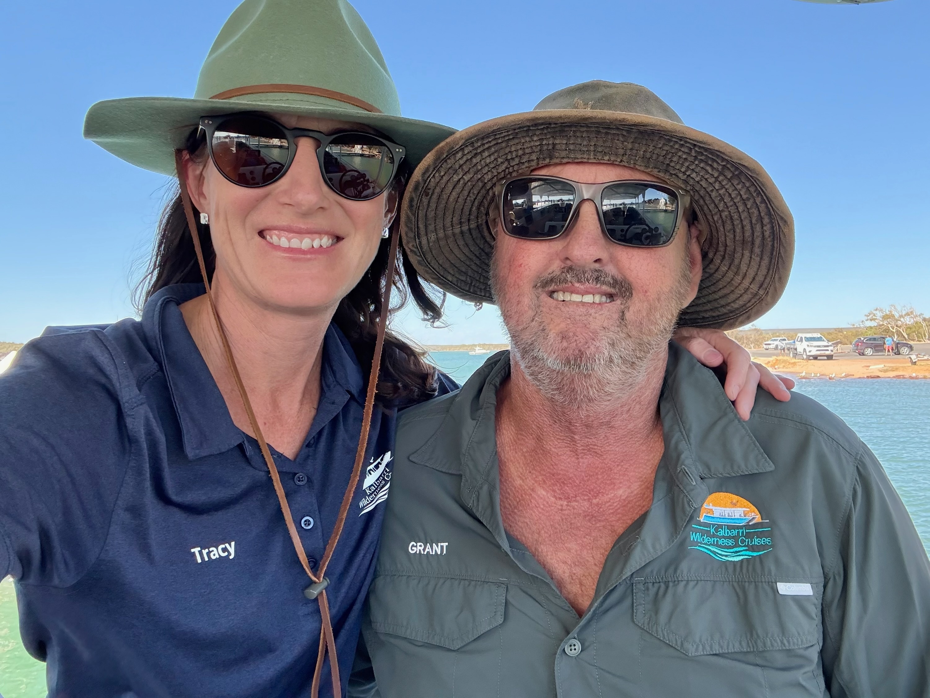 A man and a woman smiling taking a selfie, both in wide brimmed hats and sunglasses, blue water in background.