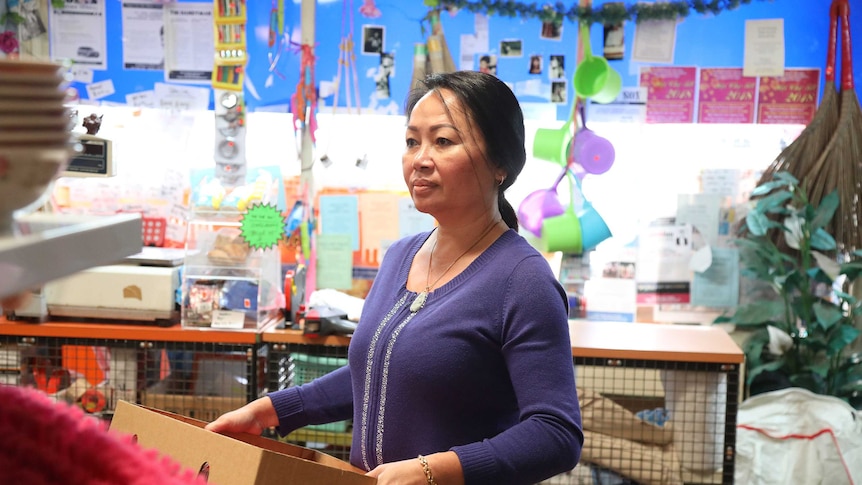 Carina Honag in character as Iris, she is in her shop, surrounded by colourful products, carrying a carboard box.