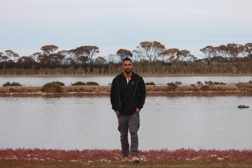 A young man walking with a lake behind him. 