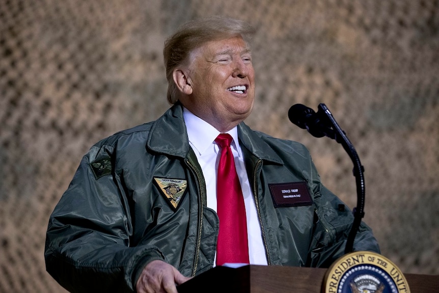 Donald Trump is in a business shirt, tie and casual jacket speaks at a podium