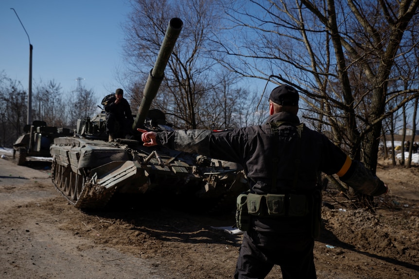 A soldier is seen from behind with his arms stretched out side to side, directing a tank