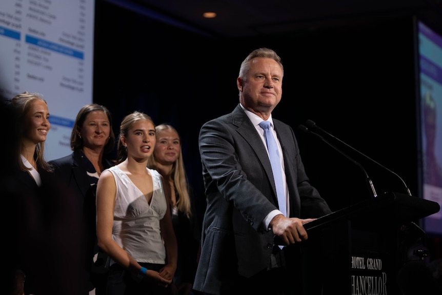 A man in a suit wearing a blue tie stands at a podium, with four women behind him.