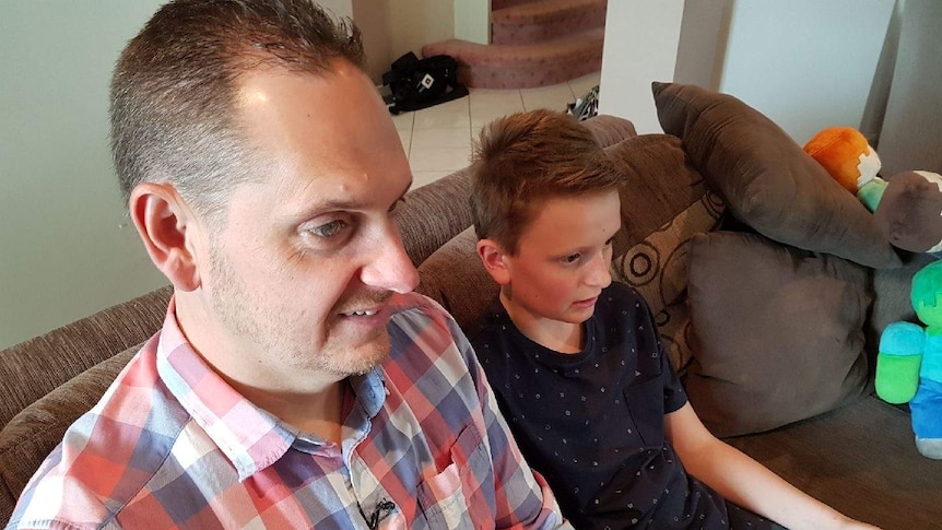 Matt Ormiston sits with his 12 year old son Joshua on a brown couch while Joshua plays a video game