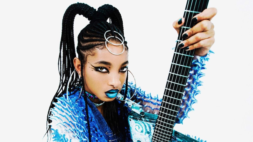 Willow smith sitting on the floor, hair braded with blue lipstick. Willow is wearing a blue spiky jumpsuit and holding a guitar.