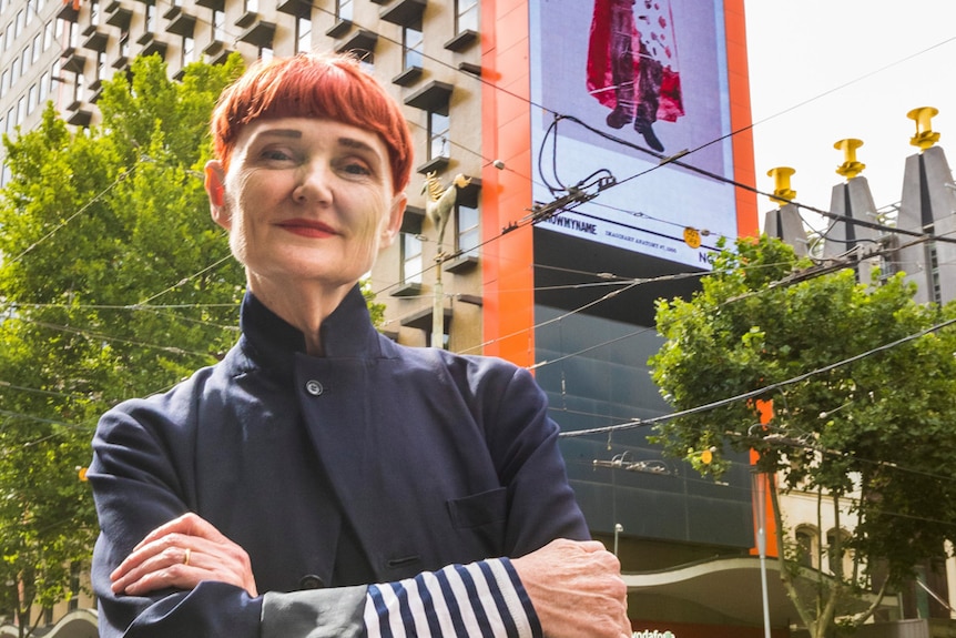 woman with short red hair standing outdoors at intersection, arms folded, looking to camera.