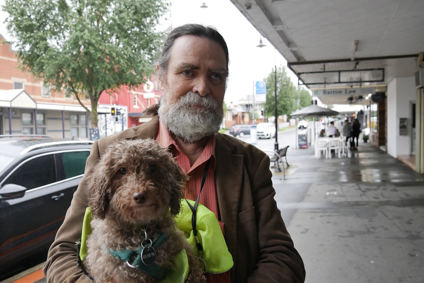 A man with a gray beard and brown jacket holds his service dog. 