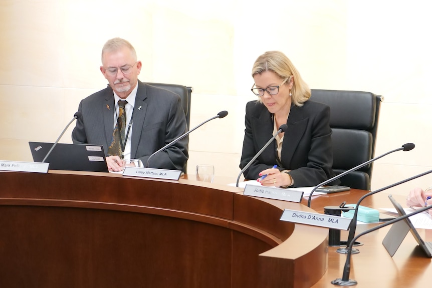 A man in a grey suit and a woman in a black suit sit at a table.