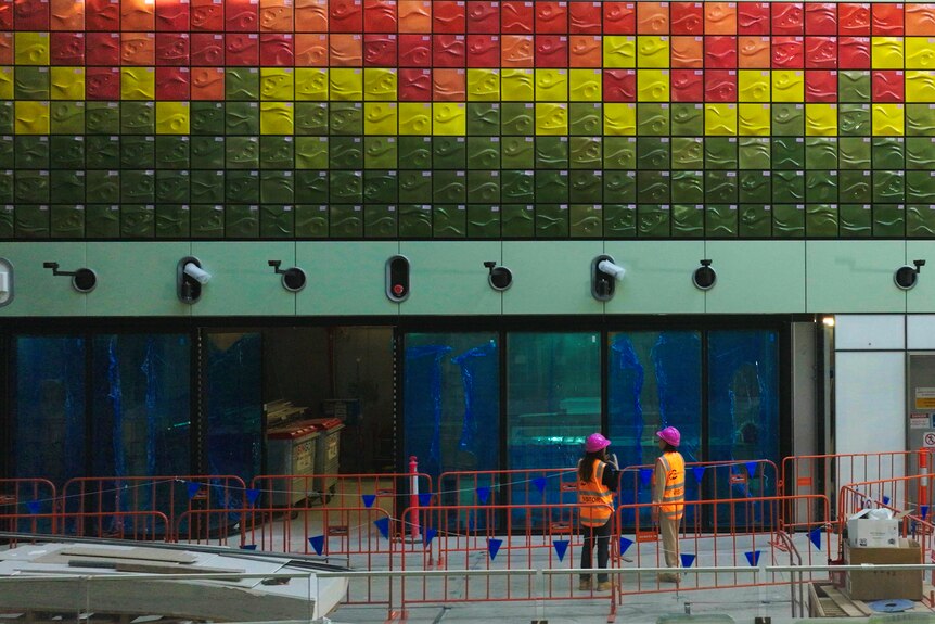 Two workers in high vis clothing and helmets look up at hundreds of colourful tiles at Parkville station