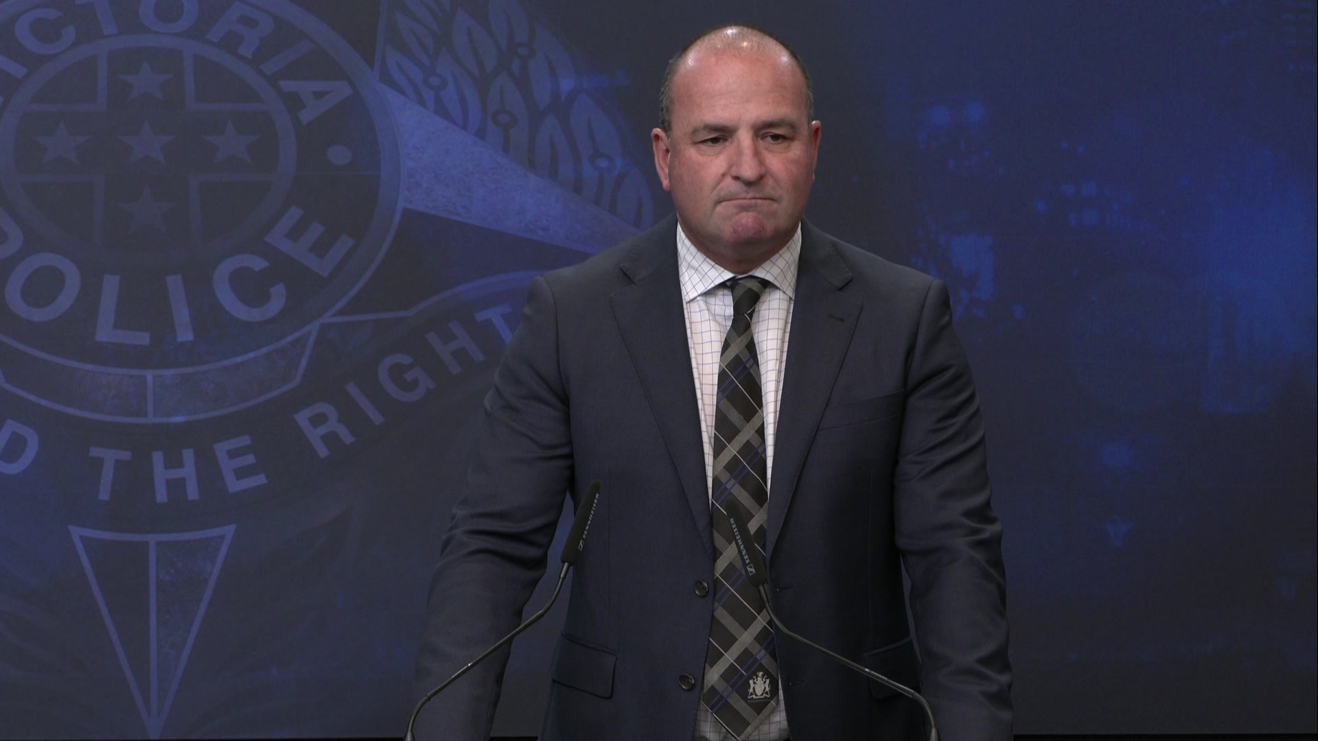 A middle-aged man in a dark suit stands in front of a Victoria Police backdrop.