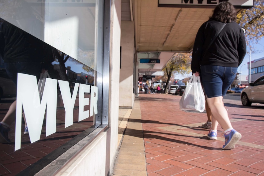 A window sign saying MYER in black and white with pedestrians walking by on the footpath