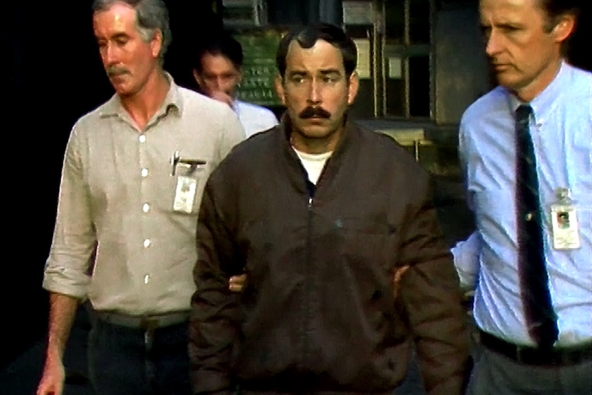A man with a moustache and brown bomber jacket being escorted by two police officers.
