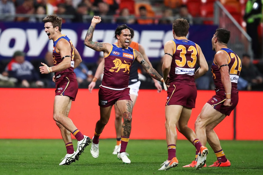 A male AFL player smiles and raises his right arm as he and three of his teammates celebrate a goal being kicked.