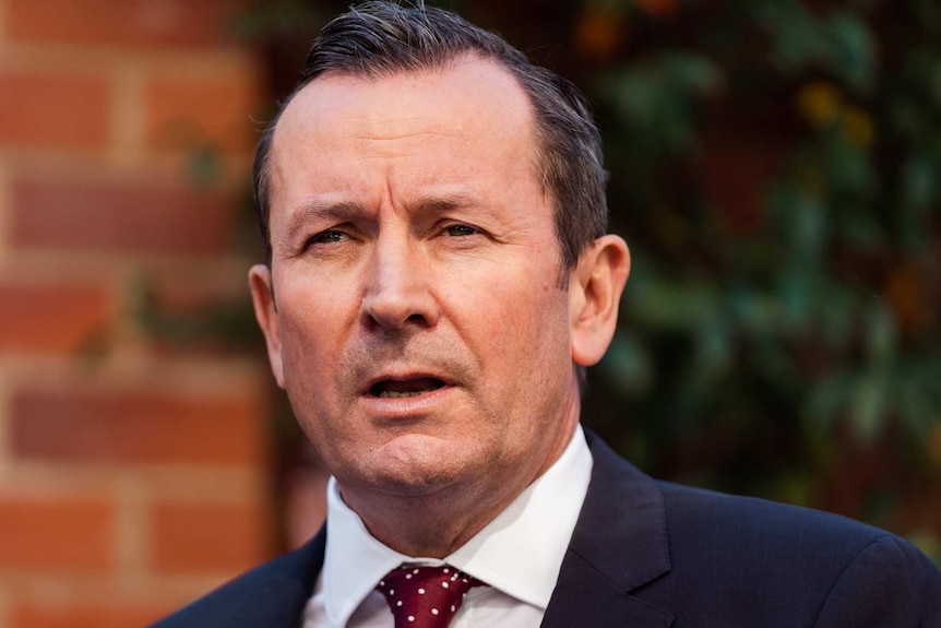 A headshot of Mark McGowan standing in a brick courtyard wearing a suit and red tie.