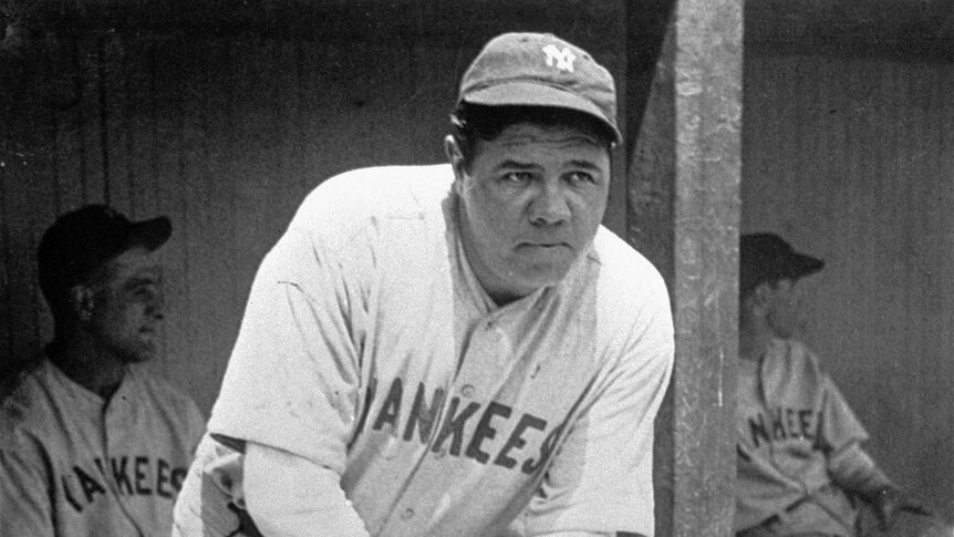 A black and white photo of Babe Ruth standing in the dugout during a baseball game in 1929