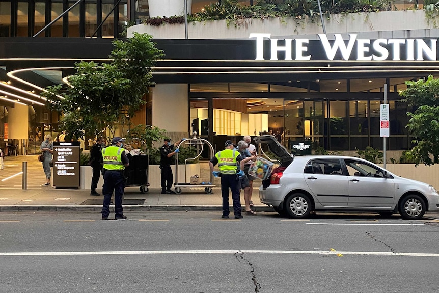Police officers watch as people put bags in a car outside hotel.