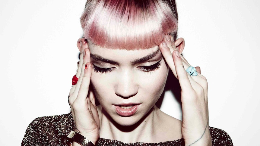 Grimes looks down with her hands on her temples. She has pink hair that has been cut short.