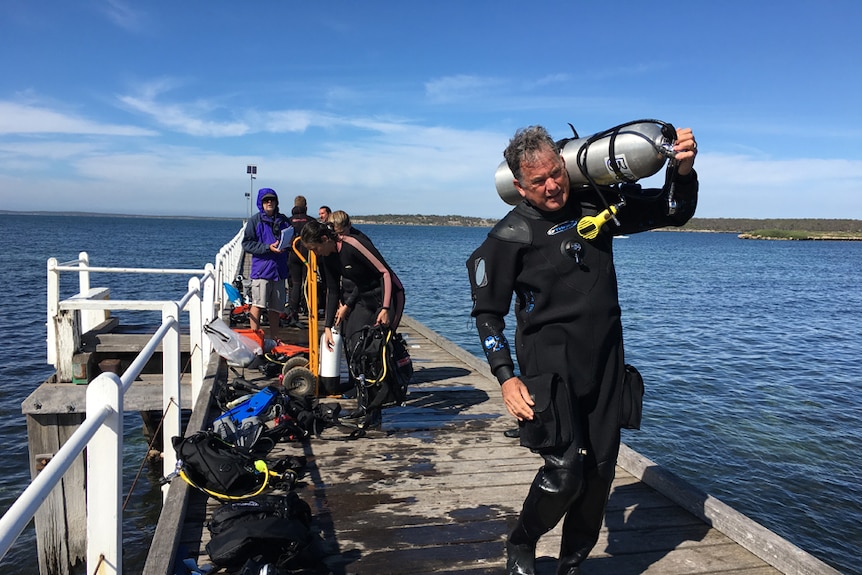 A man in a wetsuit carrying a scuba tank along a jetty with a crowd of people in the background.