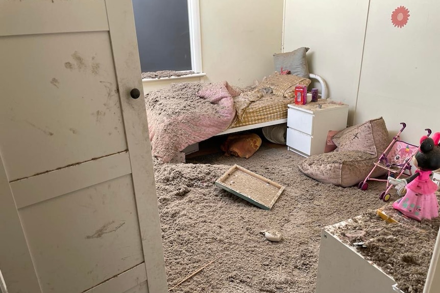 A little girls room with a pink quilt and pink toys, but it is all covered in dust and dirt that has fallen from the roof. 