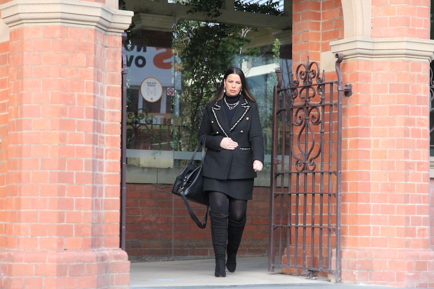 A woman wearing black clothing and sunglasses leaves a red-brick building.
