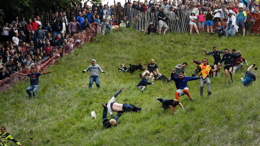 View looking up hill of people tumbling down a grassy hillside as they chase a large, round cheese, with hundreds looking on.