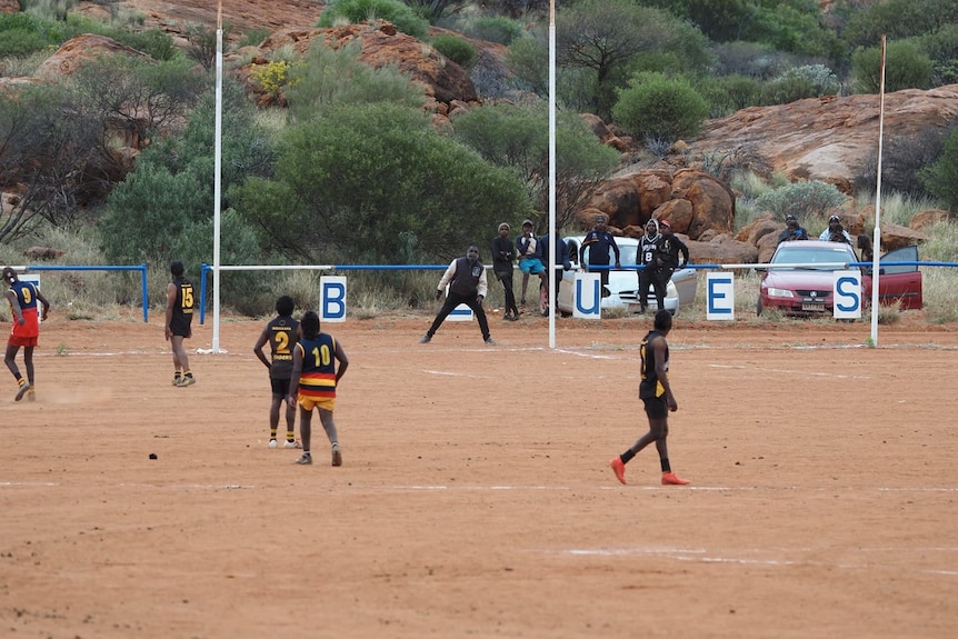 player kicking for goal with the goal umpire moving to get into position with players and spectators watching