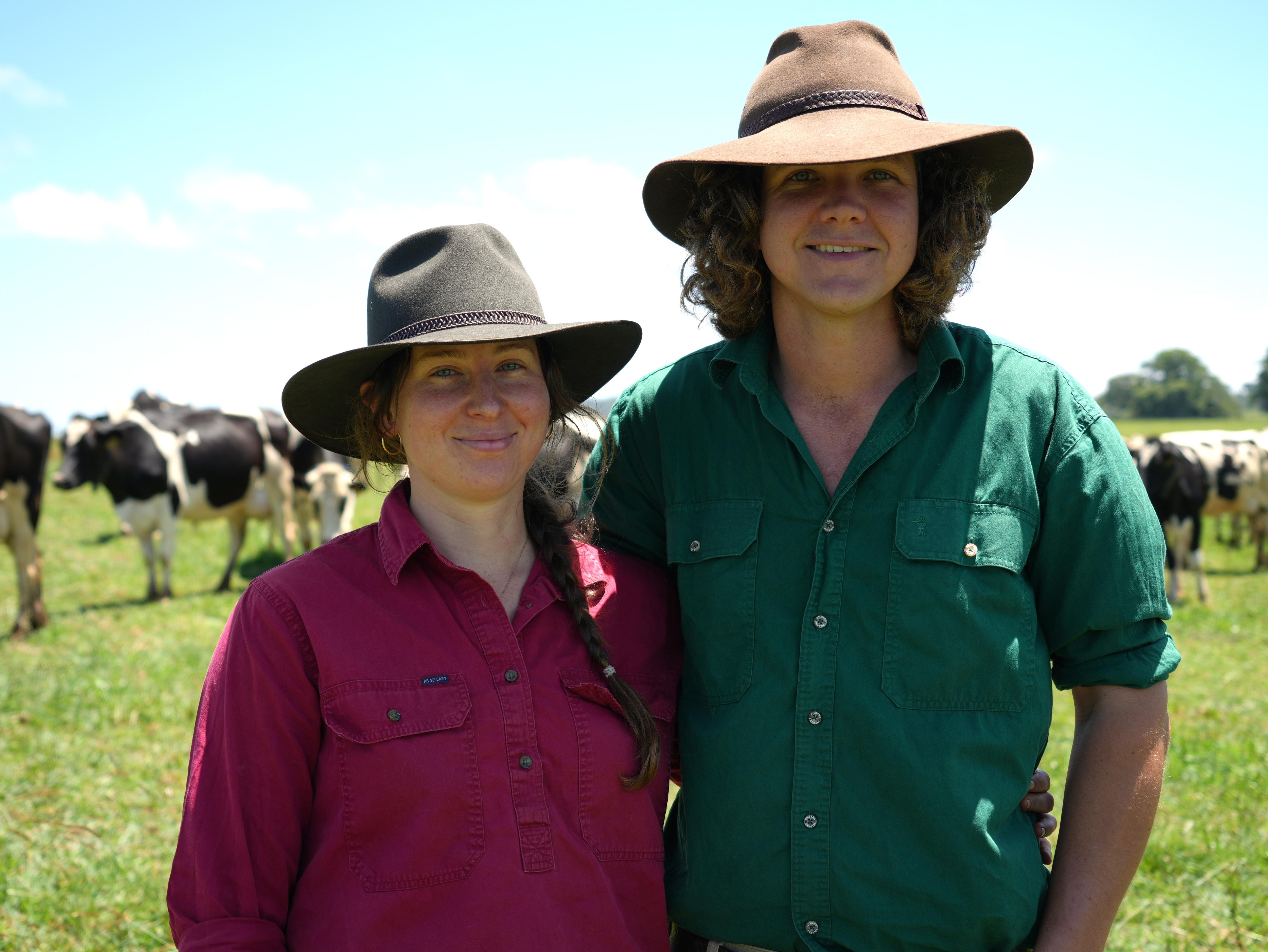 Dairy farming couple smile in the paddock with dairy cows in the background.
