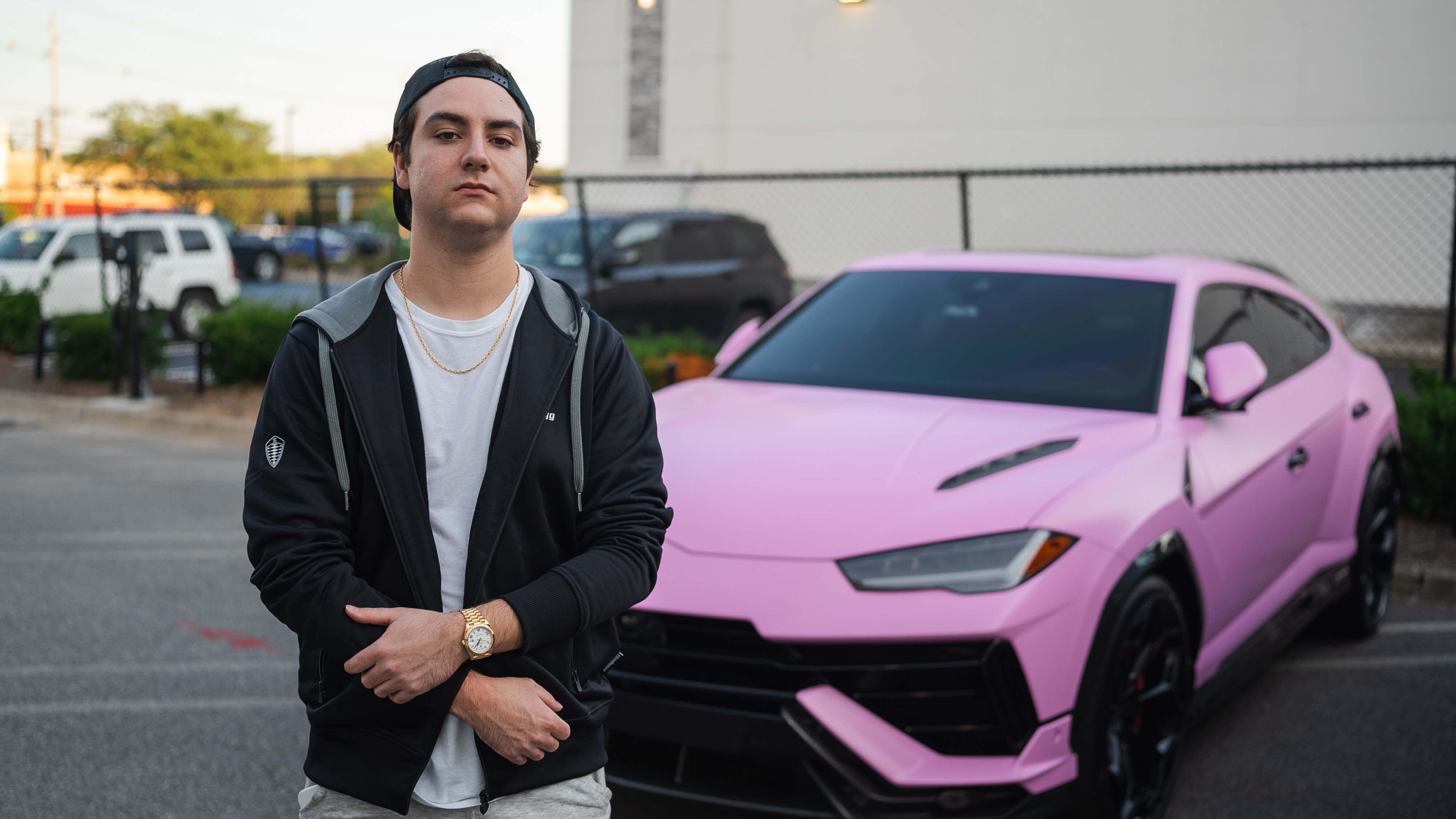 A man in his 20s wearing a white t-shirt, navy hoodie and backwards cap stands in front a pink Lamborghini in a parking lot.